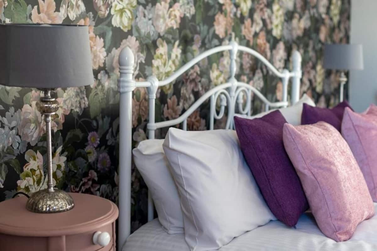 A bed with white and purple pillows, a metal headboard, and a floral wallpaper background.