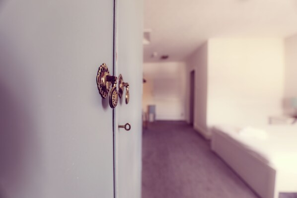 A hotel room with a bed, a door with a keyhole, and a hallway leading to another room.