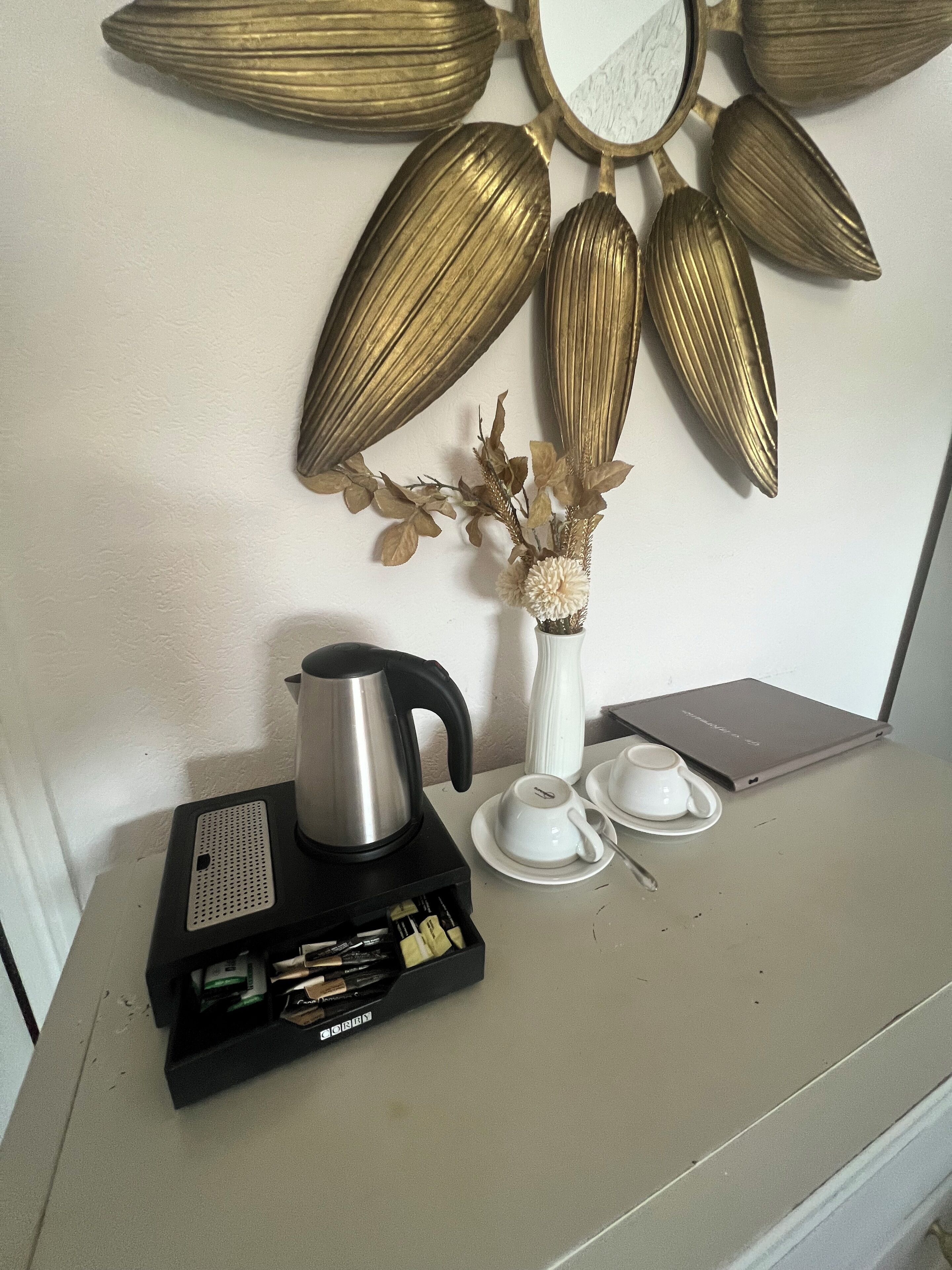 Una cafetera, un jarrón blanco con flores secas y una computadora portátil sobre una mesa.