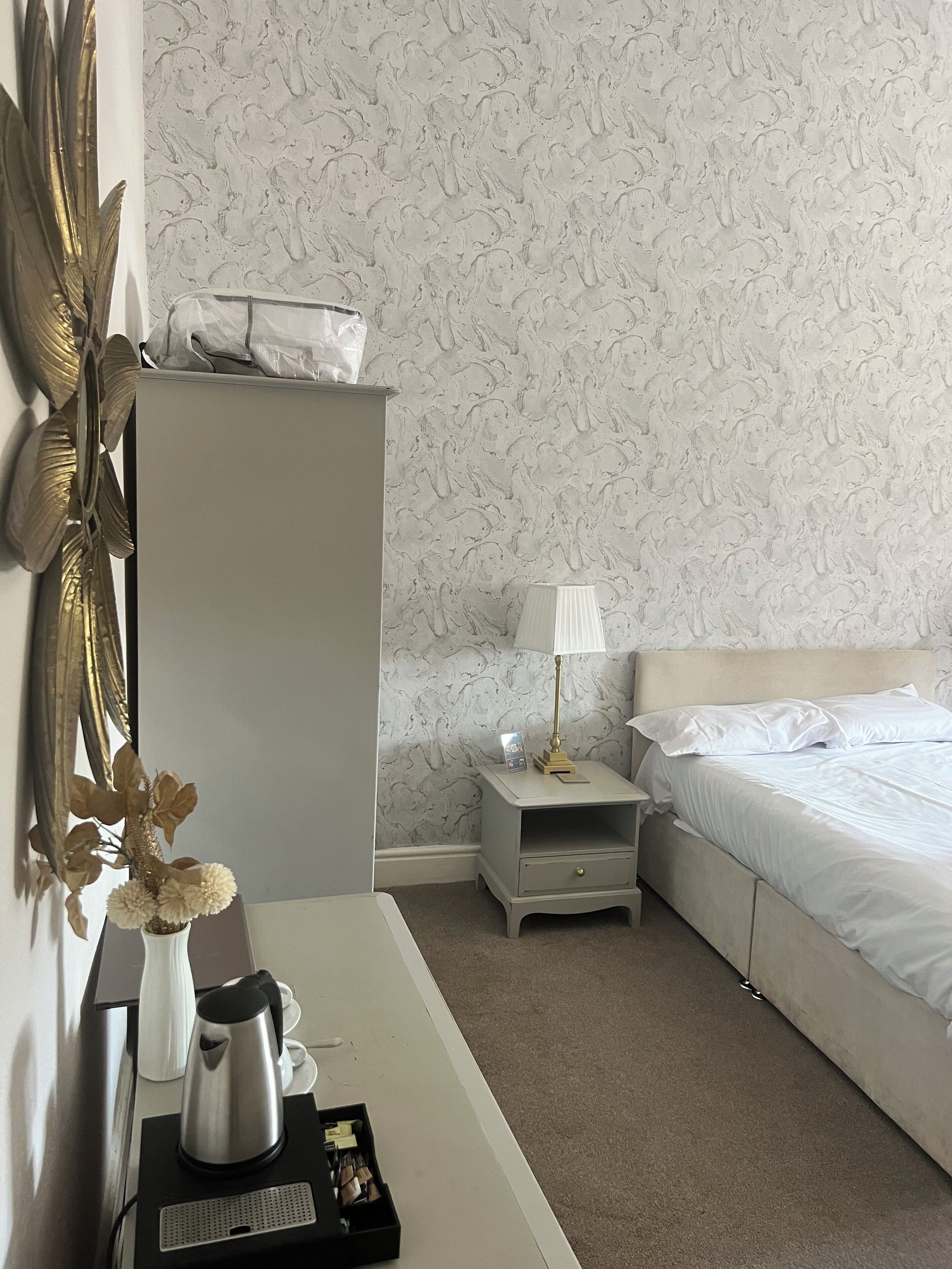 A hotel room with a bed, a nightstand with a lamp, a desk with a kettle and a small plant, and a decorative mirror.