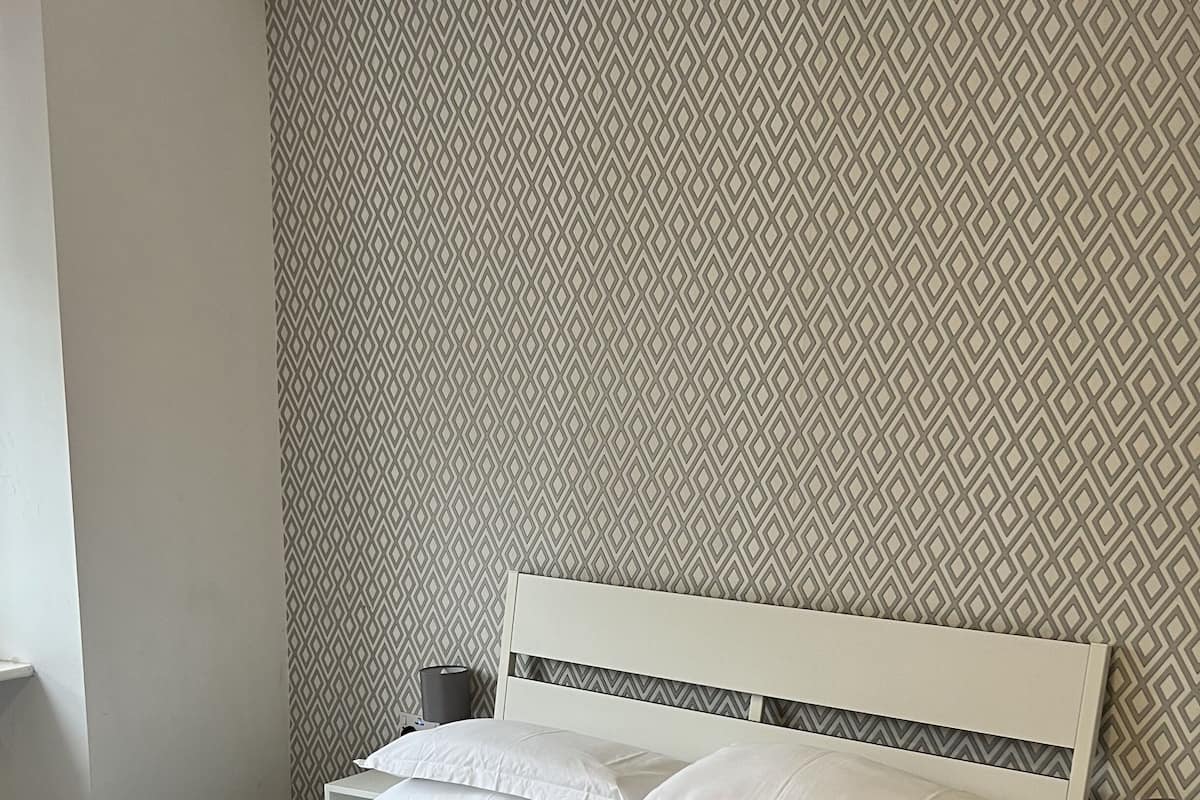 A neatly made bed with white linens and towels, a white headboard, and a patterned wallpaper.
