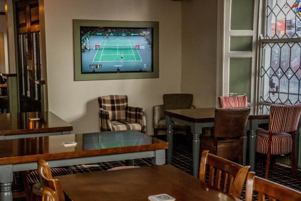 A room with a television displaying a tennis match, a plaid armchair, a wooden table, and striped chairs.
