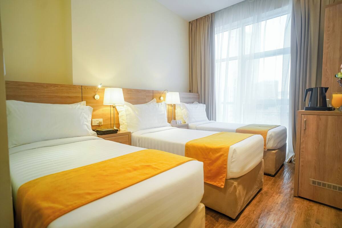 A hotel room with three beds, each with a white mattress and a yellow blanket. There are bedside lamps, a wooden headboard, and a large window with curtains.