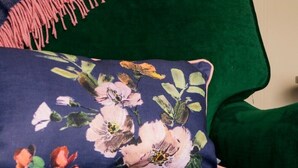 Un sofá tapizado en verde con un cojín floral y una manta azul oscuro con flecos rosas.