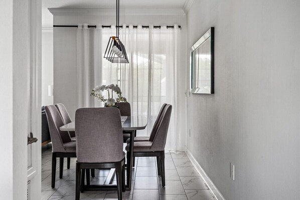 A dining area with a table and chairs, a hanging light fixture, and a window with sheer curtains. - Elegant and Cozy pool bbq (Kissimmee)