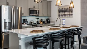 A modern kitchen with stainless steel appliances, a central island with bar stools, pendant lighting, and a large window with a view of trees.