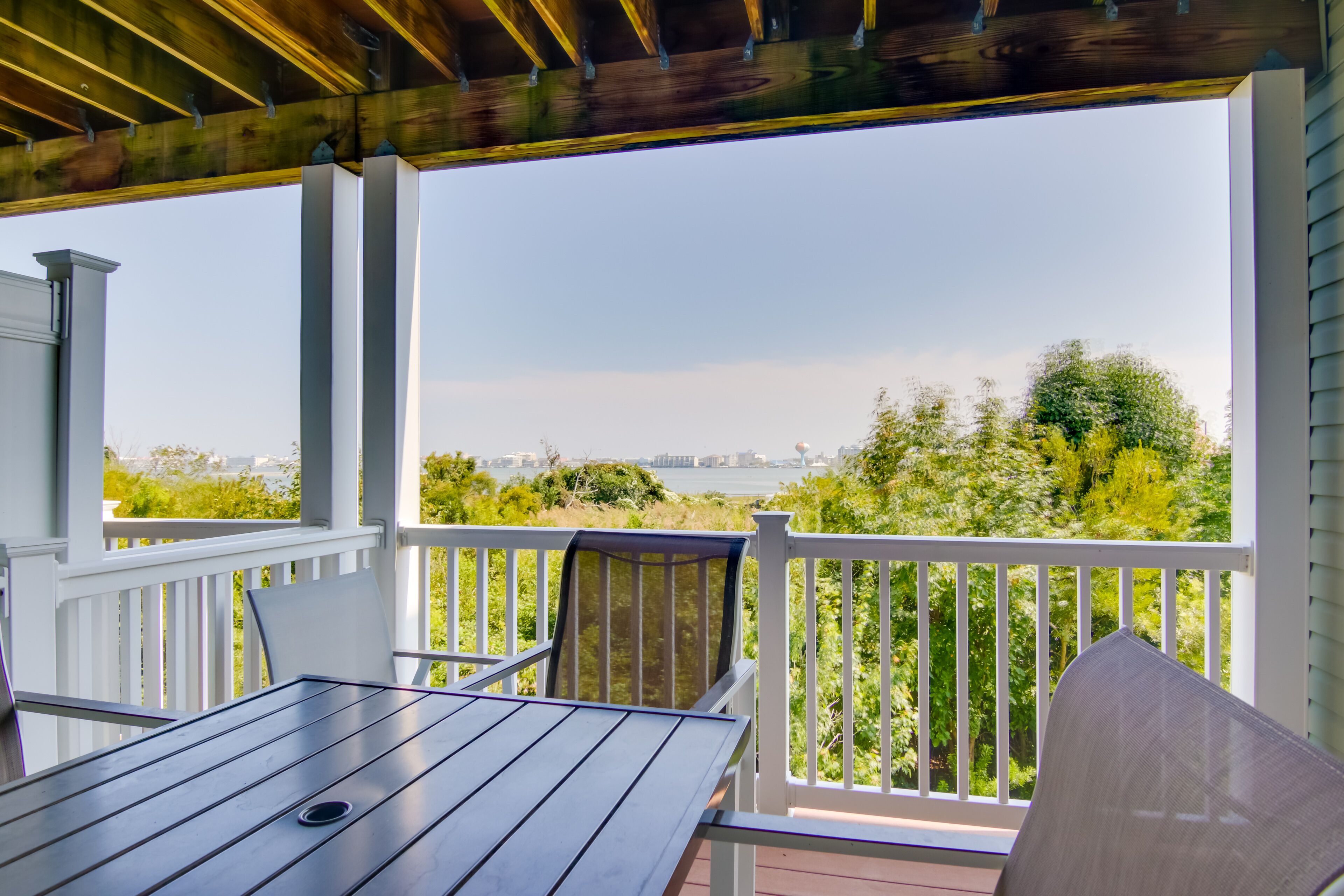 A covered outdoor area with a table and chairs, a white railing, and a view of greenery and a distant cityscape.