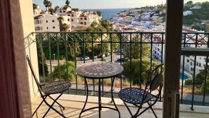 A balcony with two chairs and a table, overlooking a coastal town with white buildings and palm trees.