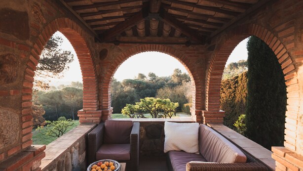 A covered outdoor seating area with brick arches, wicker furniture, and a view of a garden. - Casa Rural Can Caponet (Llica d'Amunt)
