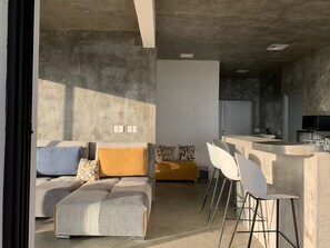 Een moderne woonkamer met een bank, voetenbankje en stoelen, een keuken met een bar, en een eettafel.