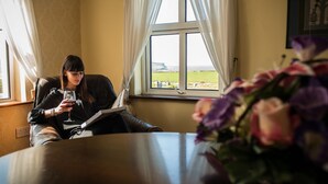 Une personne assise près d’une fenêtre, avec vue sur la mer, tenant un verre de vin et lisant un livre.
