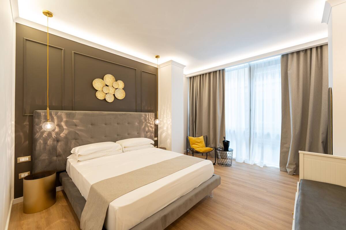 A modern hotel room with a large bed, a wall-mounted light fixture, a small side table, and a view of the outdoors through large windows.
