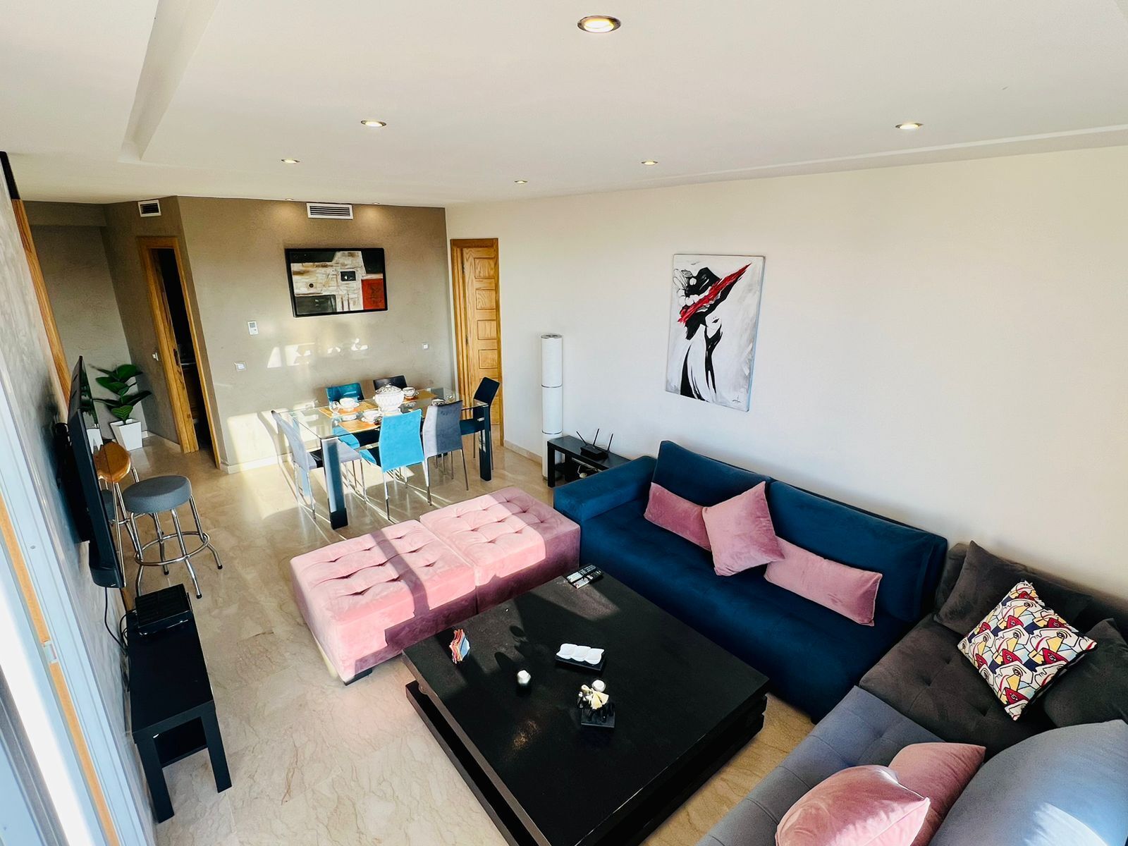 Ein modernes Wohnzimmer mit einem blauen Sofa, einem pinken Ottomanen und einem schwarzen Couchtisch. Im Hintergrund ist ein Essbereich mit Tisch und Stühlen zu sehen.