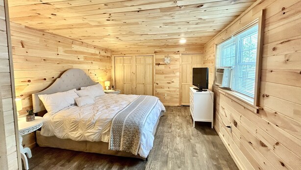 Un dormitorio con paneles de madera, una cama, mesitas de noche, un televisor y una ventana con persianas.