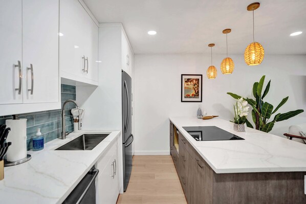 A modern kitchen with white cabinets, a black sink, and a wooden countertop. There are hanging pendant lights and a potted plant on the island.