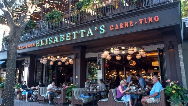 An outdoor seating area of Elisabetta's Carne Vino with tables and chairs, surrounded by plants and hanging lights.