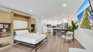 A modern hotel room with a bed, a desk, a kitchenette, and a large window with a city view.