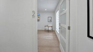 A hallway with white walls, a wooden floor, and a door with a number '2' sign. There is a framed picture on the wall and a chair in the distance.
