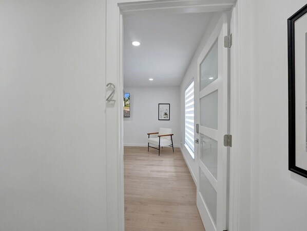 A hallway with white walls, a wooden floor, and a door with a number '2' sign. There is a framed picture on the wall and a chair in the distance.