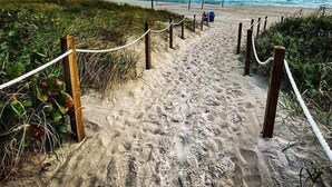 A sandy beach path with wooden posts and ropes, leading to a beach area with a lifeguard tower in the distance.