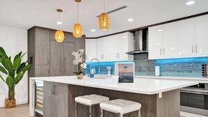 A modern kitchen with a central island, bar stools, pendant lighting, and a backsplash with blue tiles. - Villa Nicole: 4-min walk to beach, off of Atlantic (Delray Beach)