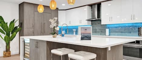 A modern kitchen with a central island, bar stools, pendant lighting, and a backsplash with blue tiles.