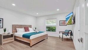 A modern bedroom with a wooden bed, a window with blinds, a TV mounted on the wall, and a chair with a blanket. - Villa Nicole: 4-min walk to beach, off of Atlantic (Delray Beach)
