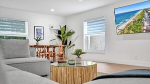 A modern living room with a grey sofa, wooden coffee table, and a large TV screen displaying a beach view. - Villa Lauren: 4-min walk to beach, off of Atlantic (Delray Beach)