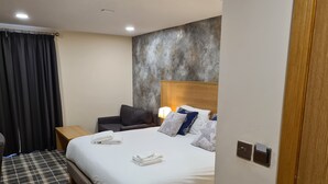 Quarto de hotel com uma cama grande, uma mesinha de cabeceira, um sofá escuro e um tapete xadrez.
