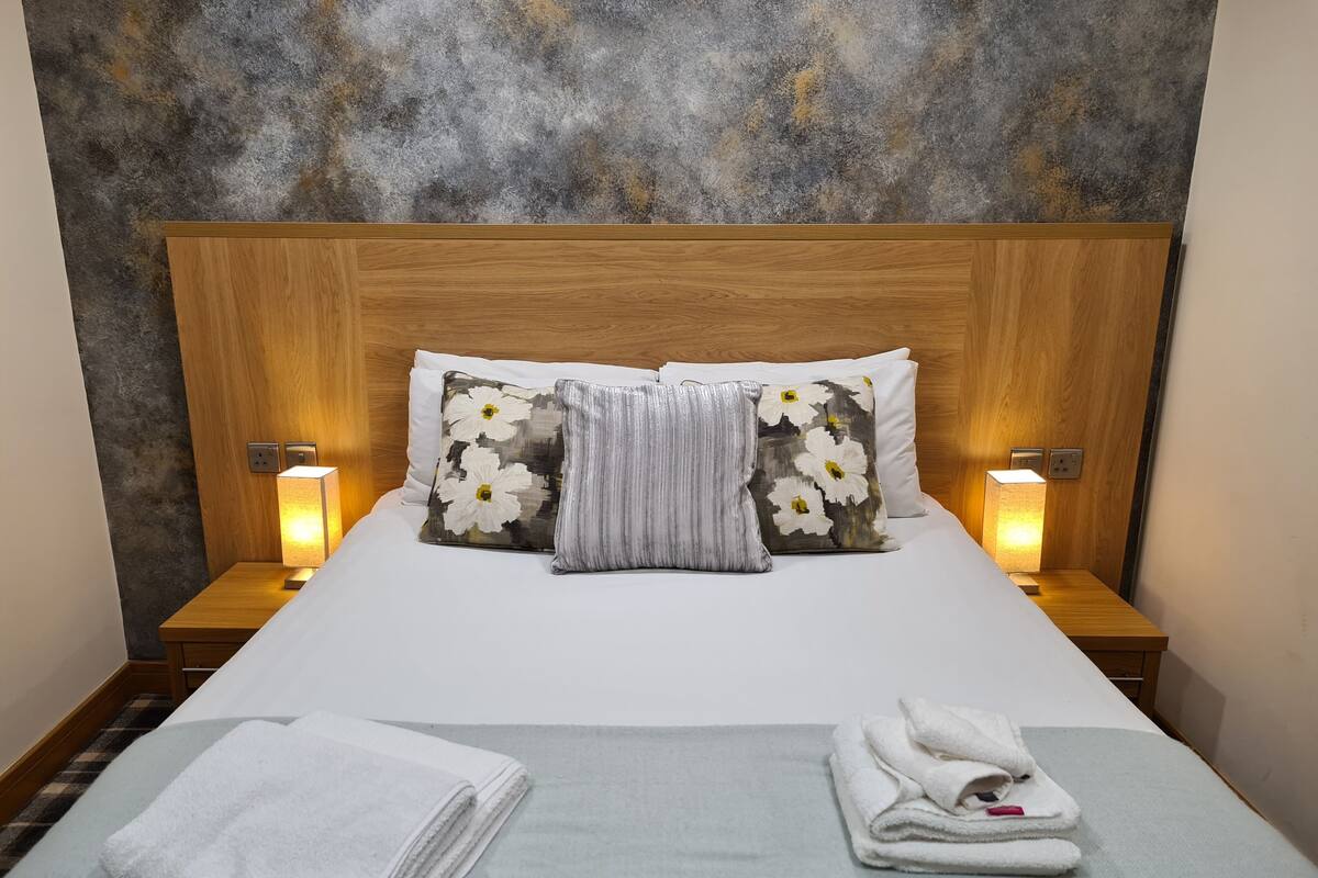 A neatly made bed with white linens, two pillows with floral designs, and a folded towel on the bed.