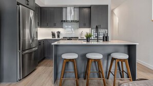 Een moderne keuken met grijze kastjes, roestvrijstalen apparatuur, een centraal eiland met krukken en een zichtbaar kookgedeelte.