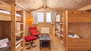 Een gezellig chaletinterieur met houten wanden, stapelbedden, een rode stoel, een kleine tafel en een raam met gordijnen.