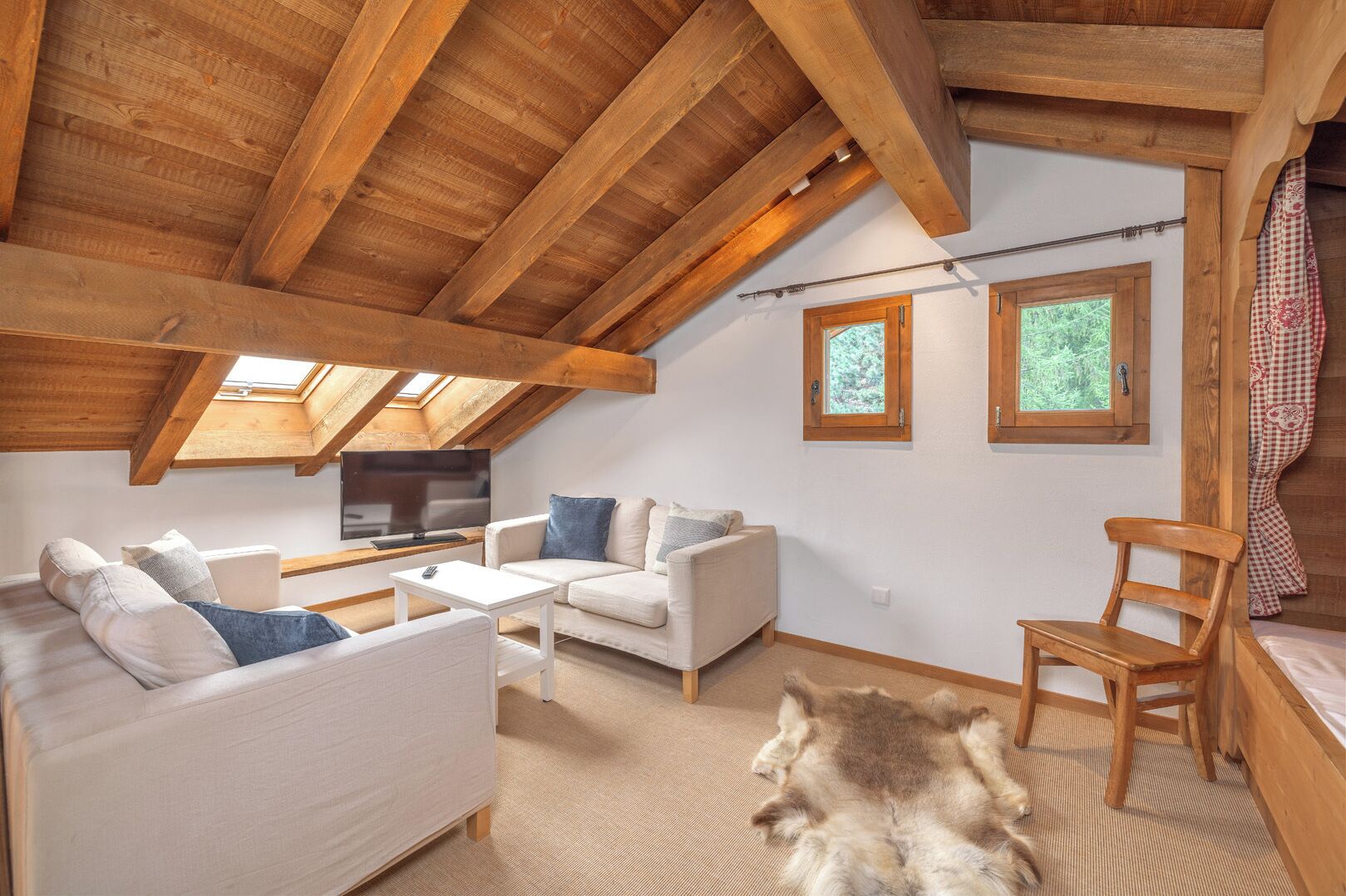 A cozy loft bedroom with a sloped ceiling, wooden beams, a large window, a television, a sofa, a chair, and a fur rug.