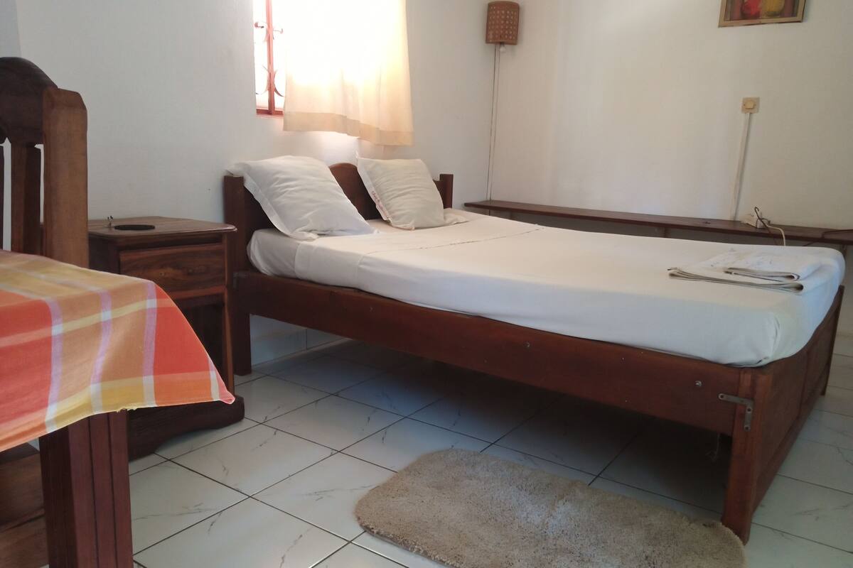 A single-bed room with a wooden frame, white bedding, a small bedside table, a nightstand, a chair, a window with sheer curtains, and a wall-mounted lamp.