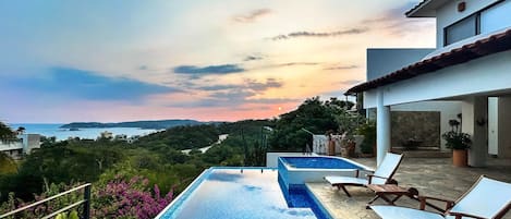 A swimming pool with lounge chairs, a stone deck, and a view of the sea at sunset.