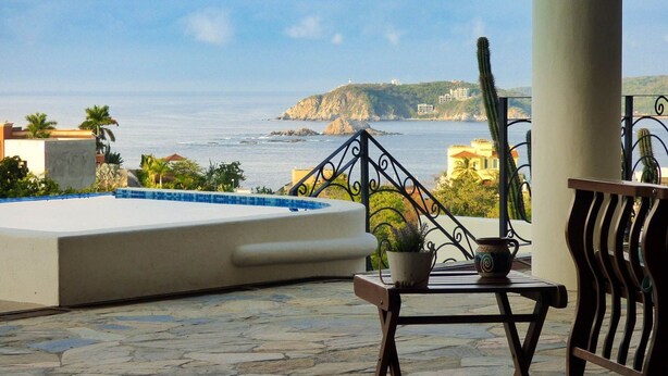 A balcony with a pool, potted plants, and a view of the sea and distant buildings. - Villa Vista Hermosa Huatulco (Santa María Huatulco)