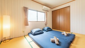 A room with a futon bed, a window with curtains, an air conditioning unit, and a wardrobe.