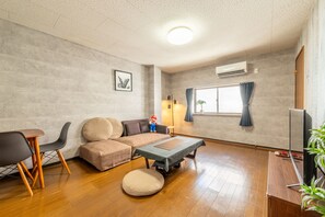 A modern living room with a sectional sofa, a small round ottoman, a coffee table, a TV stand, and a window with curtains. - DoumoTengachaya (Osaka)