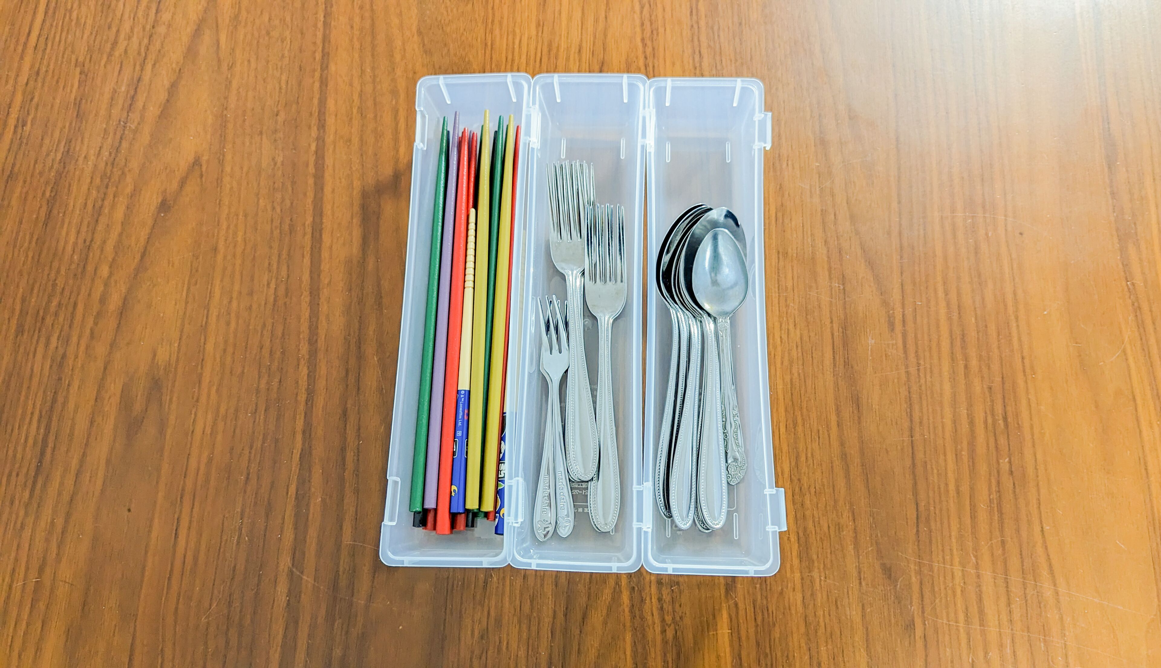 Un recipiente de plástico con cubiertos y lápices sobre una superficie de madera.