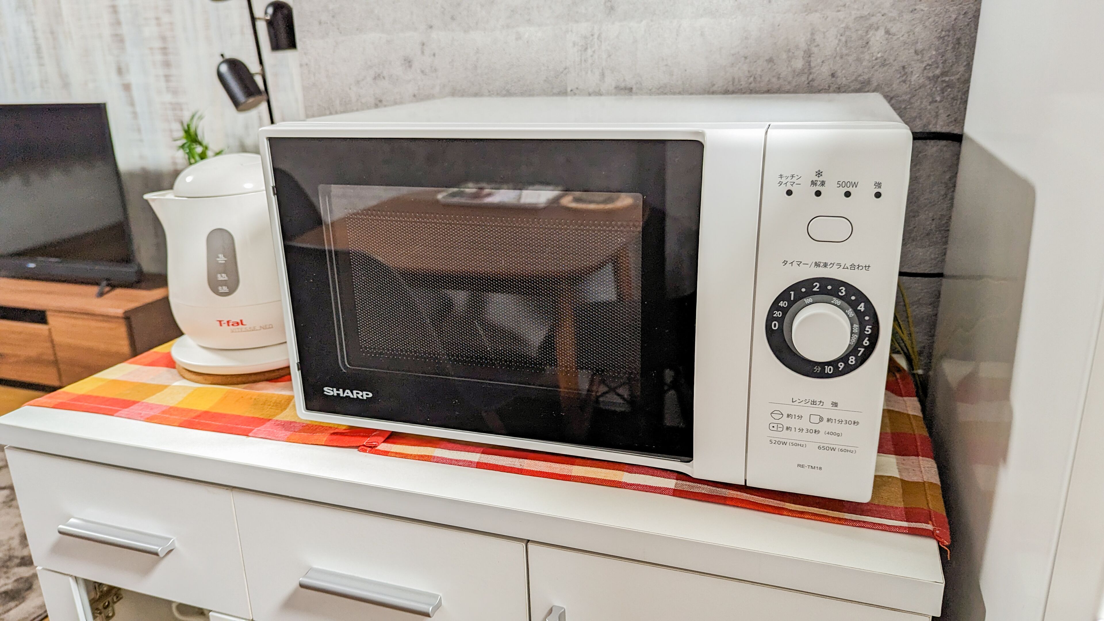 A microwave oven on a white cabinet with a red and yellow checkered cloth underneath.