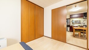A room with a wooden wardrobe, a small table, and a kitchen area visible through an open door.