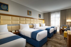 Habitación de hotel con tres camas, cabecera de madera, escritorio con lámpara y vistas al exterior a través de amplias ventanas.