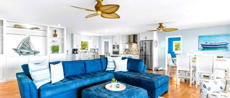 A modern living room with a blue sectional sofa, a matching ottoman, and a dining area with white chairs.