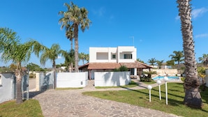 Una casa moderna de dos pisos con un camino de piedra que lleva a un área de piscina, rodeada de palmeras y un cielo azul despejado.