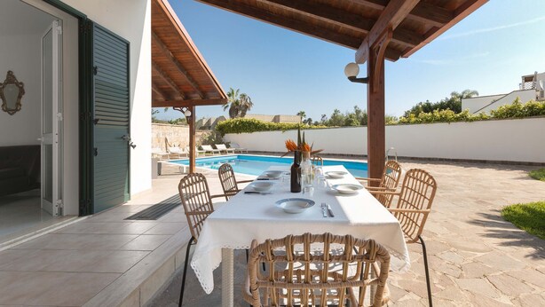 Un área de comedor al aire libre con una mesa dispuesta para cuatro personas, rodeada de sillas de mimbre, bajo una pérgola de madera, con una piscina al fondo.