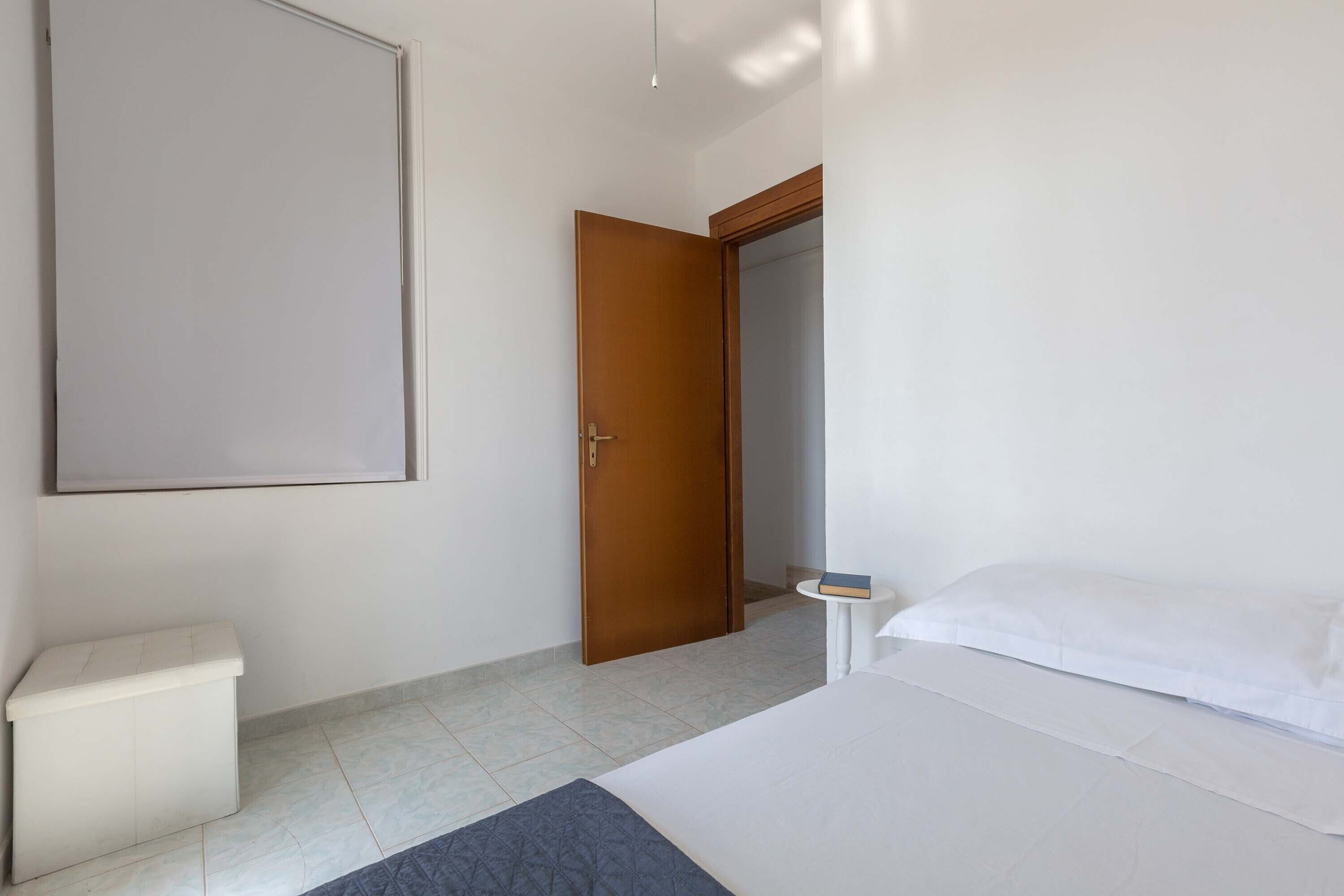 A bedroom with a bed, a bedside table, a nightstand, a wardrobe, and a door leading to another room.