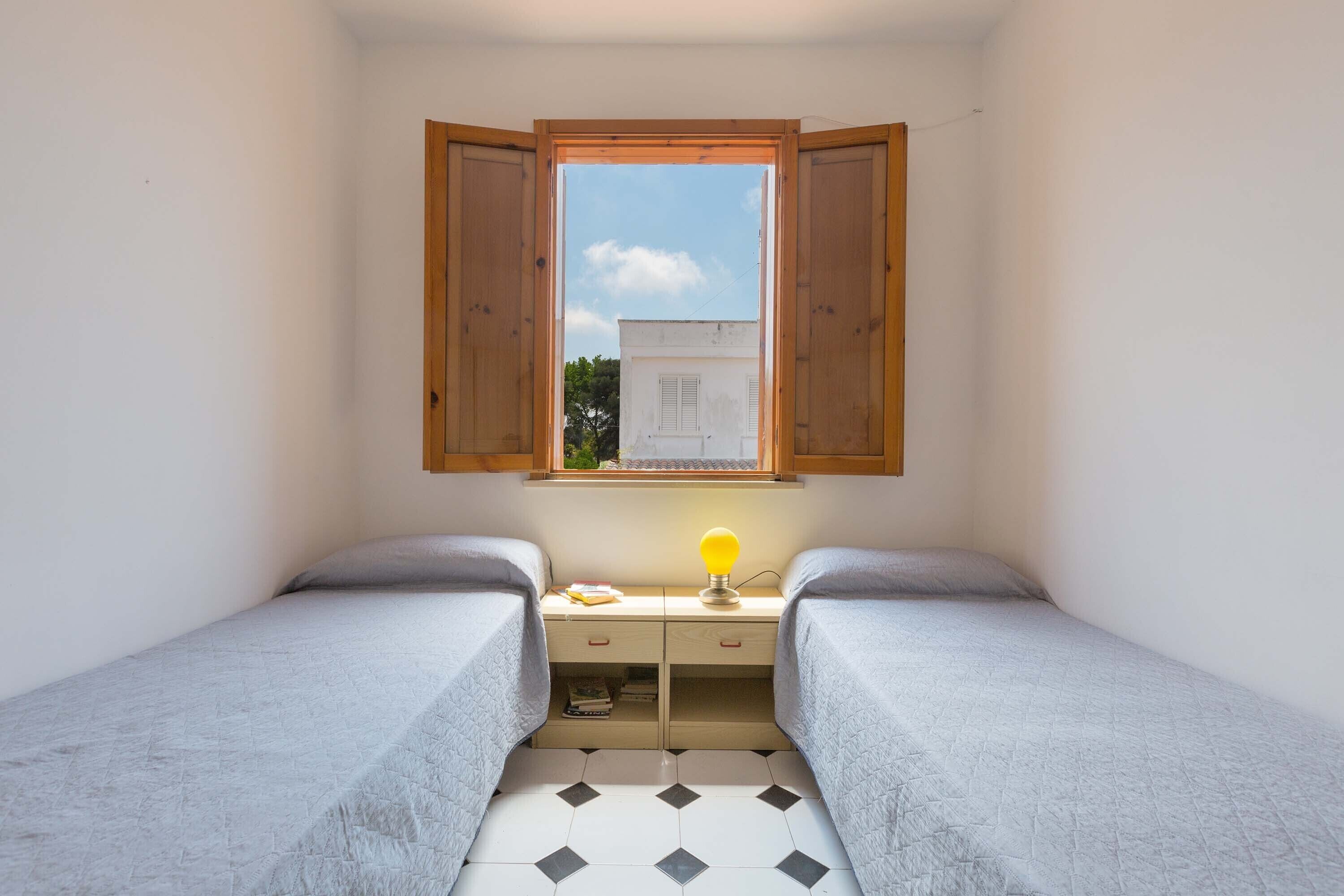 A small bedroom with two single beds, a window with wooden shutters, a bedside table with a lamp, and a tiled floor.
