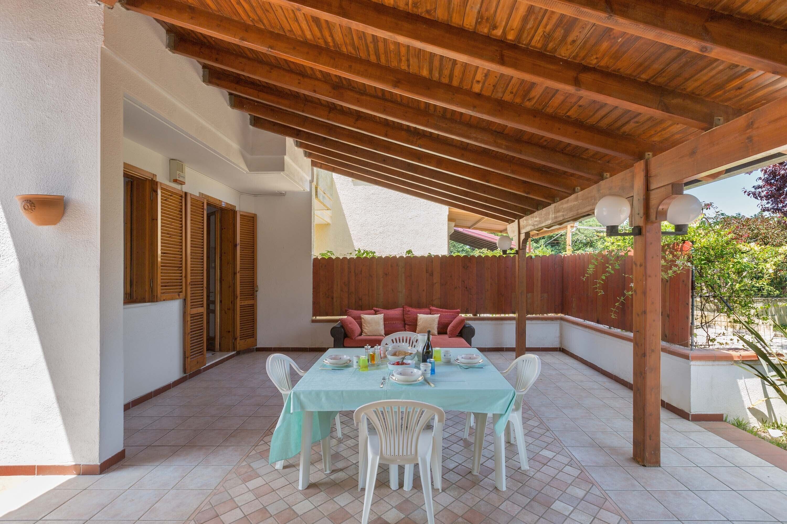 A covered patio with a wooden ceiling, a dining table set with plates and glasses, and a sofa with cushions.