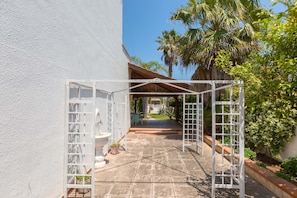 Un espace extérieur abrité, doté d’une structure métallique blanche, d’un sol en pierre et offrant une vue sur un jardin planté de palmiers.