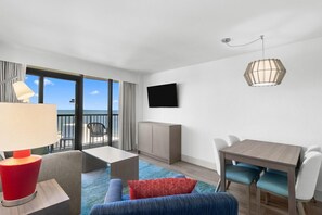A hotel room with a balcony, a flat-screen TV, a dining table with chairs, a sofa, and a lamp.